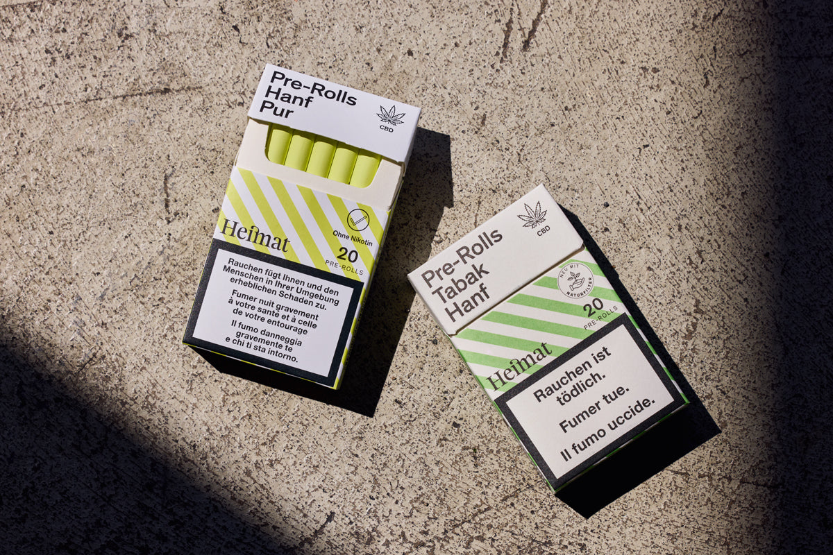 Zwei Packungen „Heimat“ Pre-Rolls liegen auf Beton: links „Hanf Pur“ mit gelbem Filter, rechts „Tabak Hanf“ mit grünem Design.