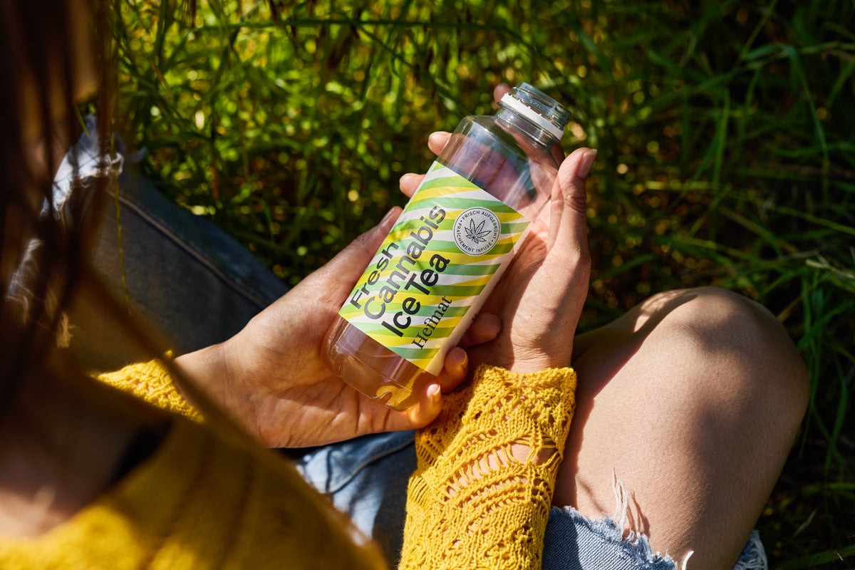 Frau mit gelbem Häkelpullover sitzt im Gras und hält eine Flasche „Fresh Cannabis Ice Tea“ von Heimat in den Händen.