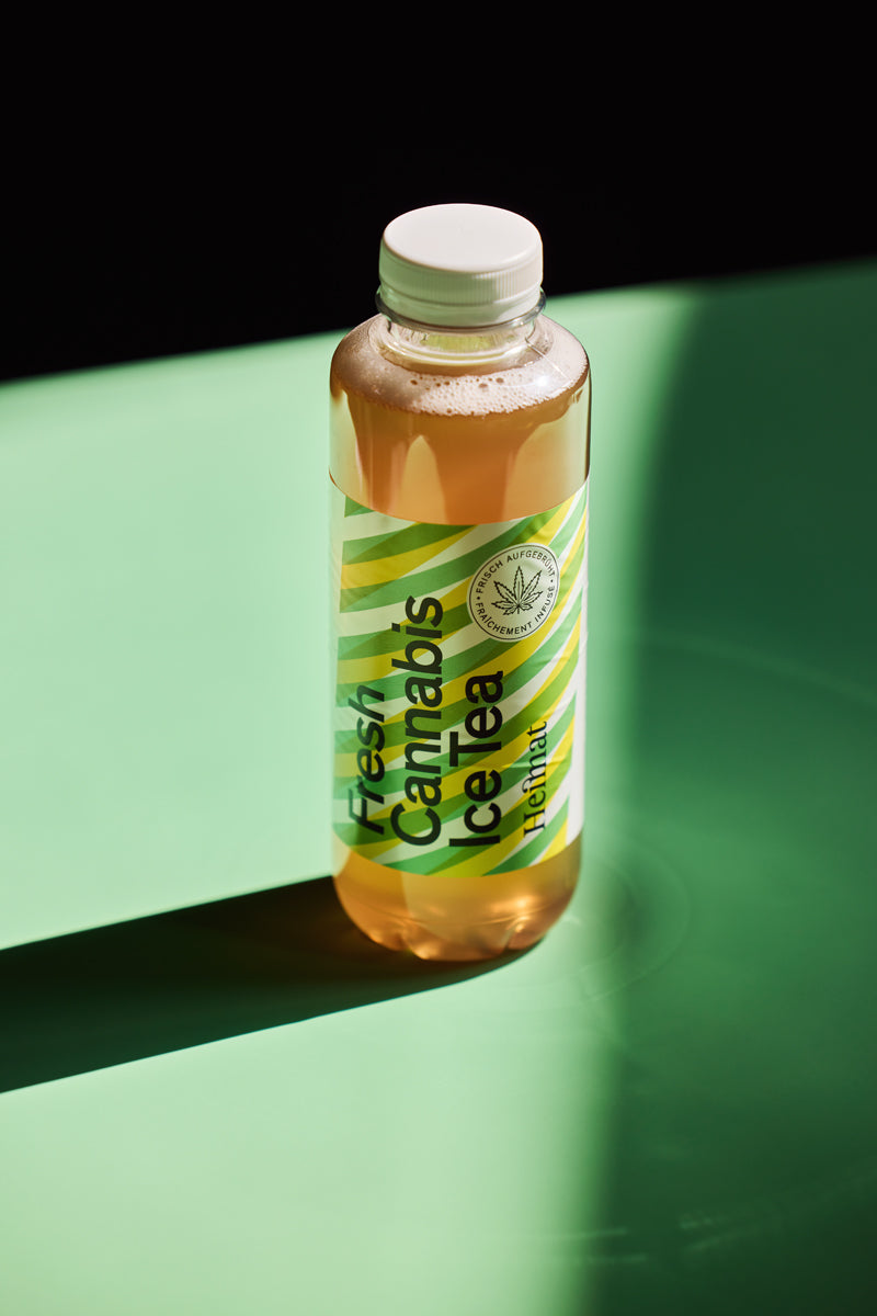 Eine Flasche „Fresh Cannabis Ice Tea“ von Heimat steht im Licht auf einem grünen Untergrund, der einen starken Schatten wirft.